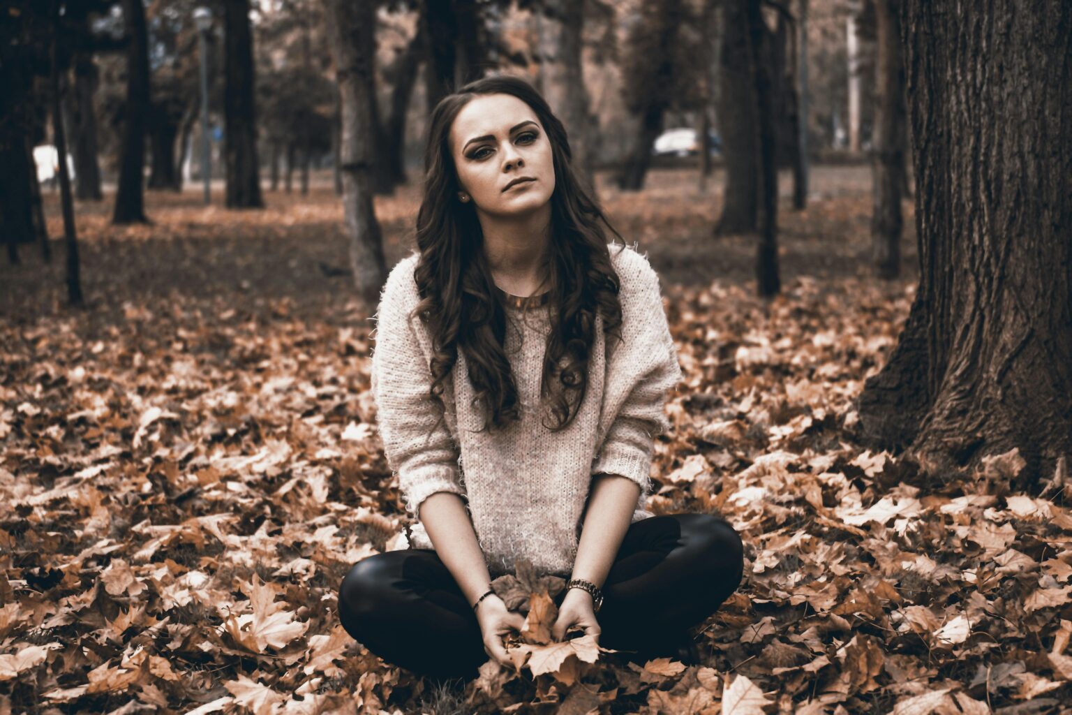 A woman sits thoughtfully on fallen leaves in a park, capturing an autumnal mood.